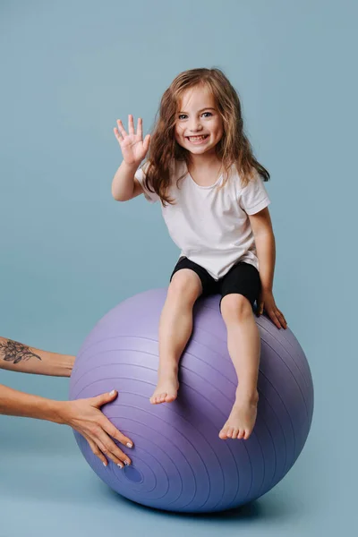 Girl sitting on a lilac fitness ball and waving a hand. — Stock Photo, Image