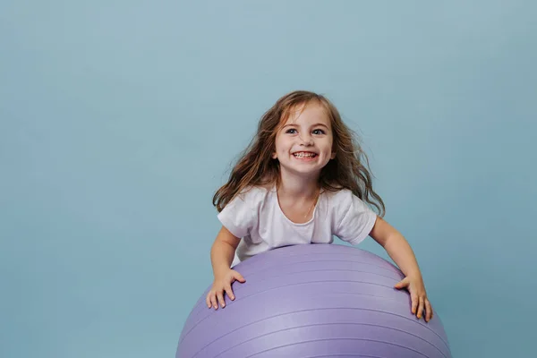 Blue-eyed curly toddler girl laughs playing on lilac fitness ball. — Stock Photo, Image