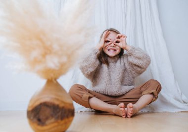 Küçük kız kelebek yoga pozisyonunda oturuyor, surat asıyor, parmak gözlüğü yapıyor.