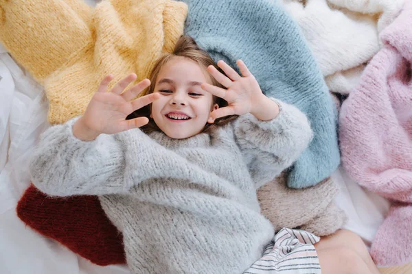 Little girl looking at her hands and fingers while lying on a pile of sweaters — Stock Photo, Image