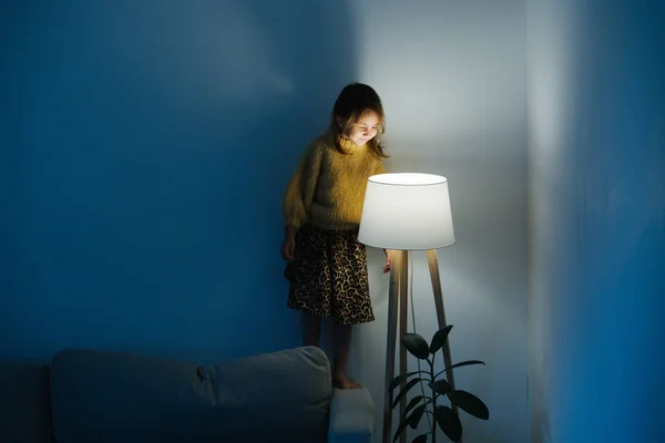 Kleines Mädchen im Pullover steht auf einer Couch und betrachtet das nächtliche Licht zu Hause. — Stockfoto