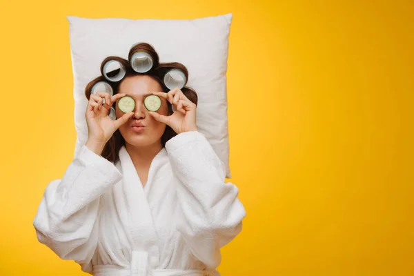 Femme au foyer sur un oreiller avec des cheveux en rouleaux tenant des tranches de concombre sur ses yeux — Photo