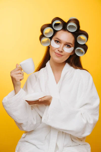 Housewife drinking tea with hair in rollers and patches under eyes over yellow — Stock Photo, Image