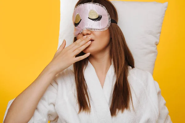 Brunette femme se couche sur l'oreiller avec masque de sommeil sur les yeux, bâillant . — Photo