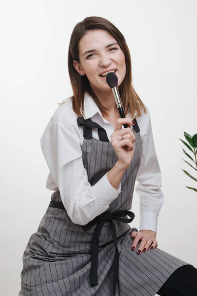 Mulher estilista em avental com braços cruzados segurando escova de maquiagem ao lado de seus lábios — Fotografia de Stock