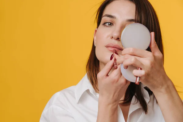 Mulher atraente em uma camisa de vestido branco segurando espelho de mão, aplicando batom — Fotografia de Stock