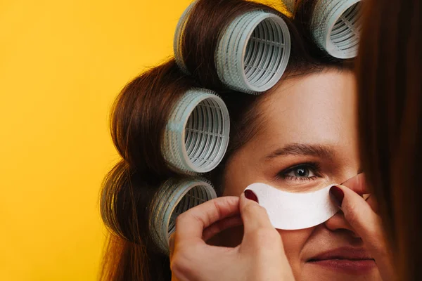 Beautician εφαρμογή μάτι-patch σε μια γυναίκα με μαλλιά σε κυλίνδρους. Κλείσιμο — Φωτογραφία Αρχείου