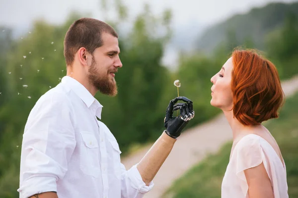 Un uomo che tiene il dente di leone in una mano bionica, per farci soffiare sopra la sua ragazza. Vista laterale . — Foto Stock