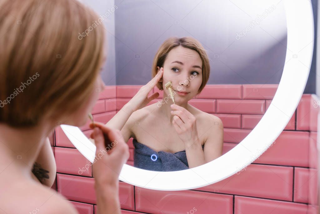 Young woman massaging her face with gua sha spiked metal roller in her bathroom