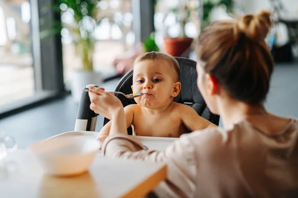 Matka krmí dítě lžící, zatímco dítě sedí na vysoké židli — Stock fotografie