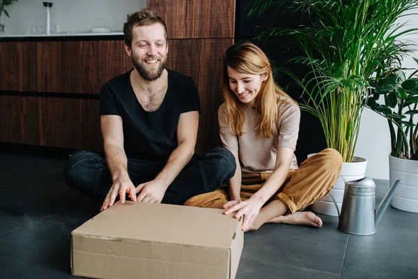 Feliz pareja moderna sentada en un piso con piernas cruzadas, caja de apertura . — Foto de Stock