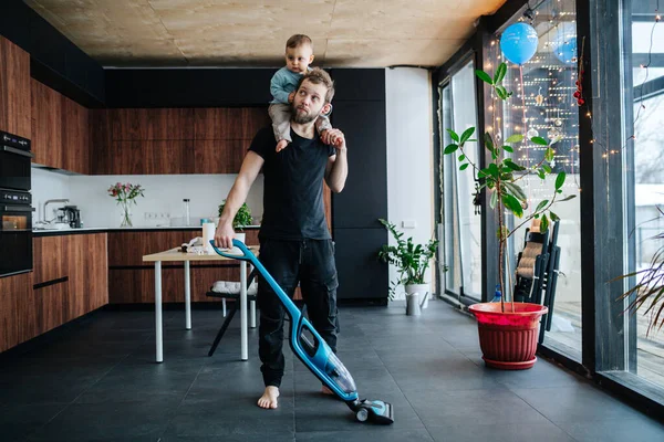 El joven padre aspira el piso del apartamento con su bebé montado en su cuello — Foto de Stock