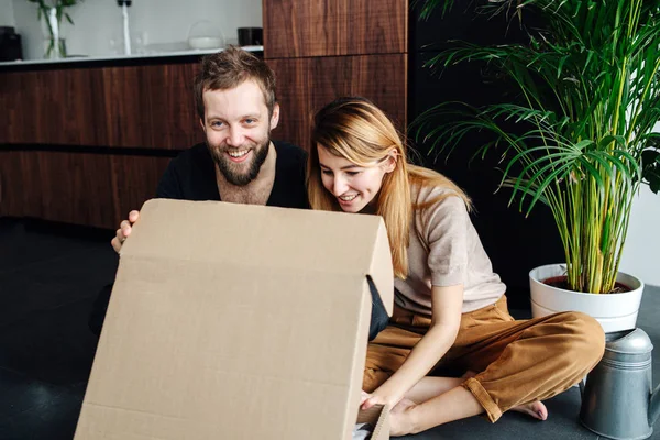 Feliz pareja moderna sentada en un piso con piernas cruzadas, caja de apertura . — Foto de Stock