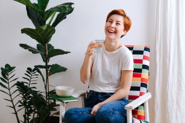 Happy middle aged woman with short ginger hair eating sitting in a chair at home with a glass of water in hands. She's wearing casual clothes: white T-shirt and jeans.