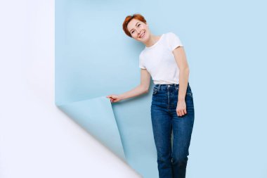 Emotionless focused middle aged woman with short ginger hair pulling piece of blue covering from a white wall. She's wearing casual clothes: white T-shirt and jeans.