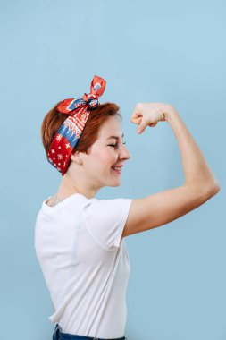 Portrait of a cheerful middle aged housewife with short ginger hair and colorful headband tencing biceps, showing off her strength. She's wearing casual clothes: white T-shirt and jeans. Over blue