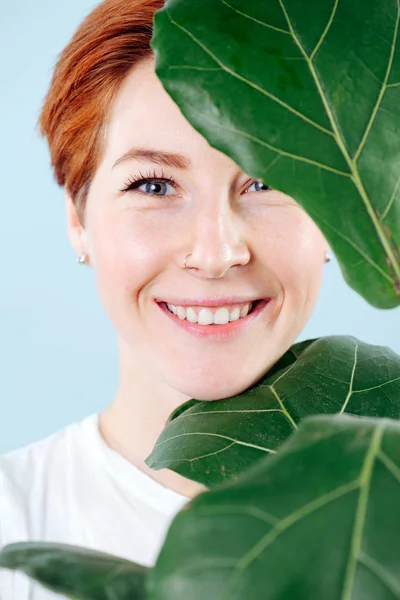 Portrait of a cheerful woman with short ginger hair behind big ficus leaves — Stock Photo, Image