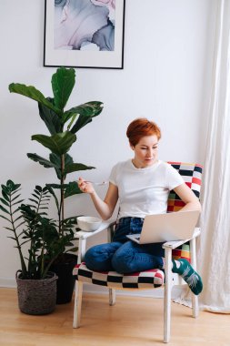 Emotionless middle aged woman with short ginger hair eating muesli in a chair at home while browsing on a chair at home. She's wearing casual clothes: white T-shirt and jeans.
