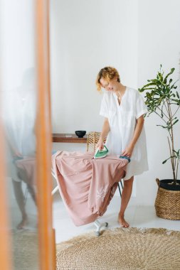 Çıplak ayaklı, neşeli, kısa sarı saçlı ve beyaz gömlekli genç bir kadın evdeki panoda giysilerini ütülüyor.