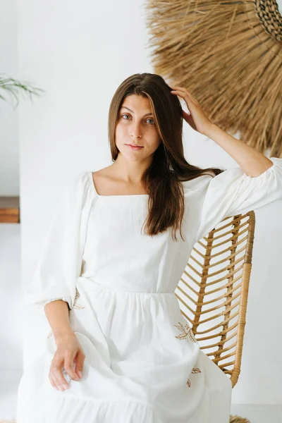 Portrait of a modest calm young woman in a light white summer dress sitting on a chair in a tropical style room. Leaning on fingers.