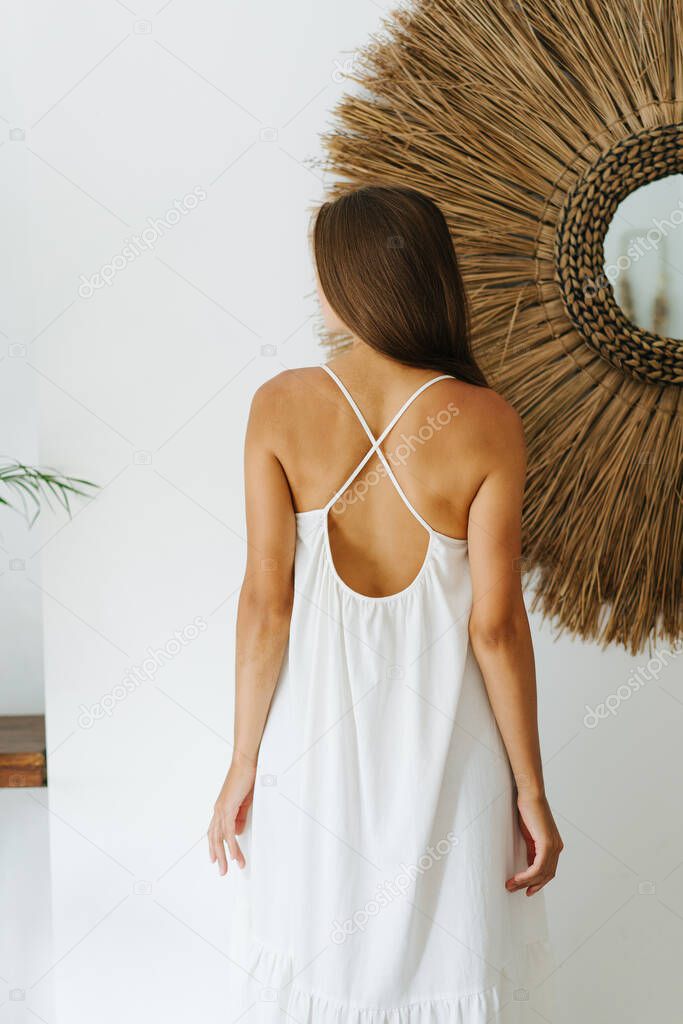 Pretty young woman in a light white summer dress posing for a photo in a tropical style room with her back turned.