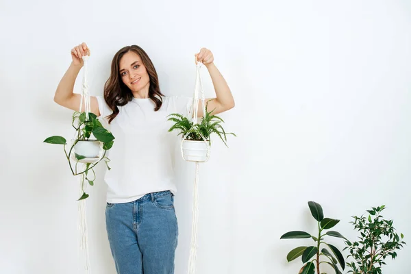 きれいな女性は美しい植物とマクラメのポットを保持しています 白い壁に白いTシャツとジーンズを着てる — ストック写真