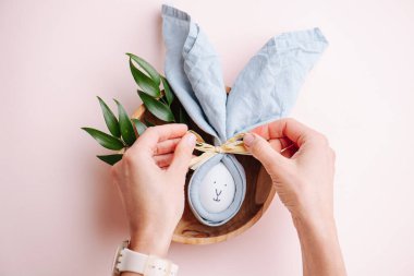 Peçetedeki paskalya yumurtasının üzerinde tavşan resmi var. Pembe kareli bir masa örtüsünün üzerindeki ahşap bir kaseye konmuş. Elleri yay bağlıyor. Üst görünüm.