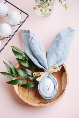 Peçetedeki paskalya yumurtasının üzerinde tavşan resmi var. Pembe kareli bir masa örtüsünün üzerindeki ahşap bir kaseye konmuş. Kavanozda taze yumurta ve papatya çiçeğinin yanında. Üst görünüm.