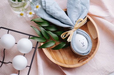 Peçetedeki paskalya yumurtasının üzerinde tavşan resmi var. Pembe kareli bir masa örtüsünün üzerindeki ahşap bir kaseye konmuş. Kavanozda taze yumurta ve papatya çiçeğinin yanında. Üst görünüm.