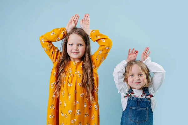 两个小女孩用双手捂着蓝色背景做着复活节兔子耳朵的手势 一个穿黄色衣服 一个穿牛仔裤工作服 — 图库照片