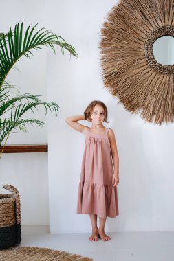 Tropikal bir odada pembe bir Midi elbiseyle poz veren, yalın ayaklı, tatlı küçük bir kız. Elleri başının arkasında..