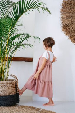 Tropikal bir odada bembeyaz giyinmiş pembe midi elbiseli, yalın ayaklı, tatlı küçük bir kız. Ayağıyla ağır saksı bitkisini itiyor. Yan görünüm.