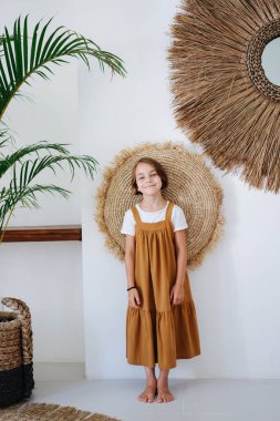 Beyaz gömleğin üzerine giyilmiş, tropikal bir odada fotoğraf için poz veren, yalın ayaklı, deniz mavisi elbiseli küçük bir kız. Sırtını yastığa yaslıyor, duvara bastırıyor..