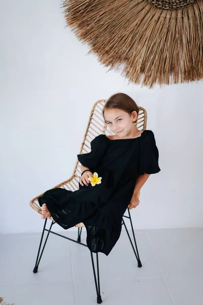 半黒のドレスを足で椅子の上に座って 熱帯のスタイルの部屋で写真のポーズをとった素敵な裸足の少女 ユリの花を持ってる — ストック写真