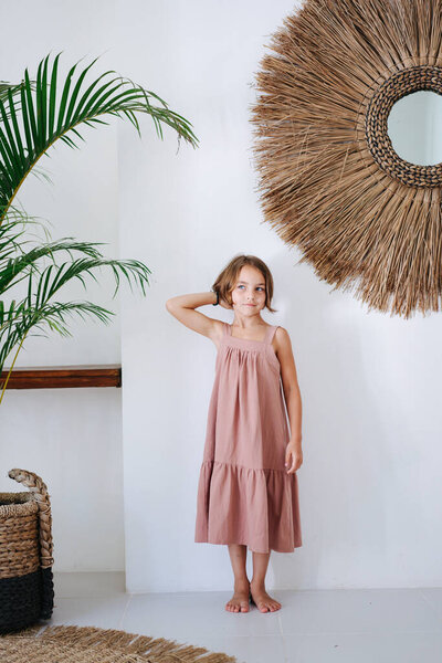 Cute barefoot little girl in a pink midi dress posing for a photo in a tropical style room. Her hand on a back of her head.