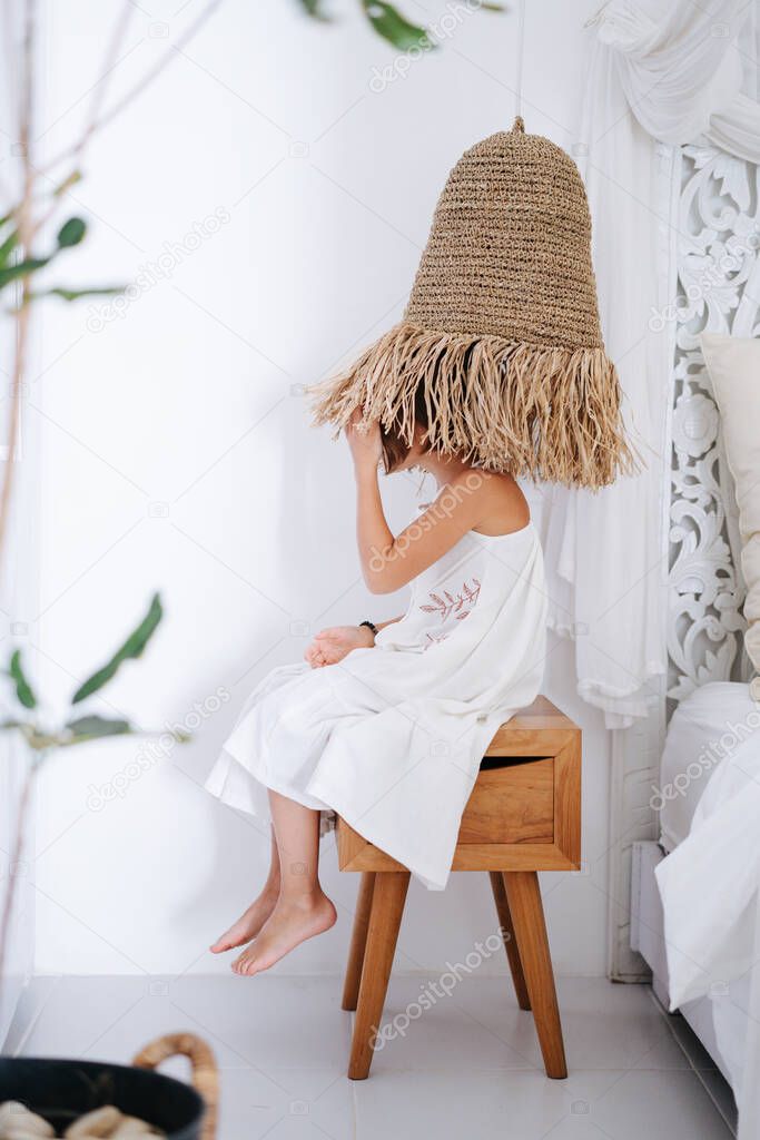 Playful little girl in a midi white dress sitting on a bedside table, wearing lampshade as a hat. Side view.