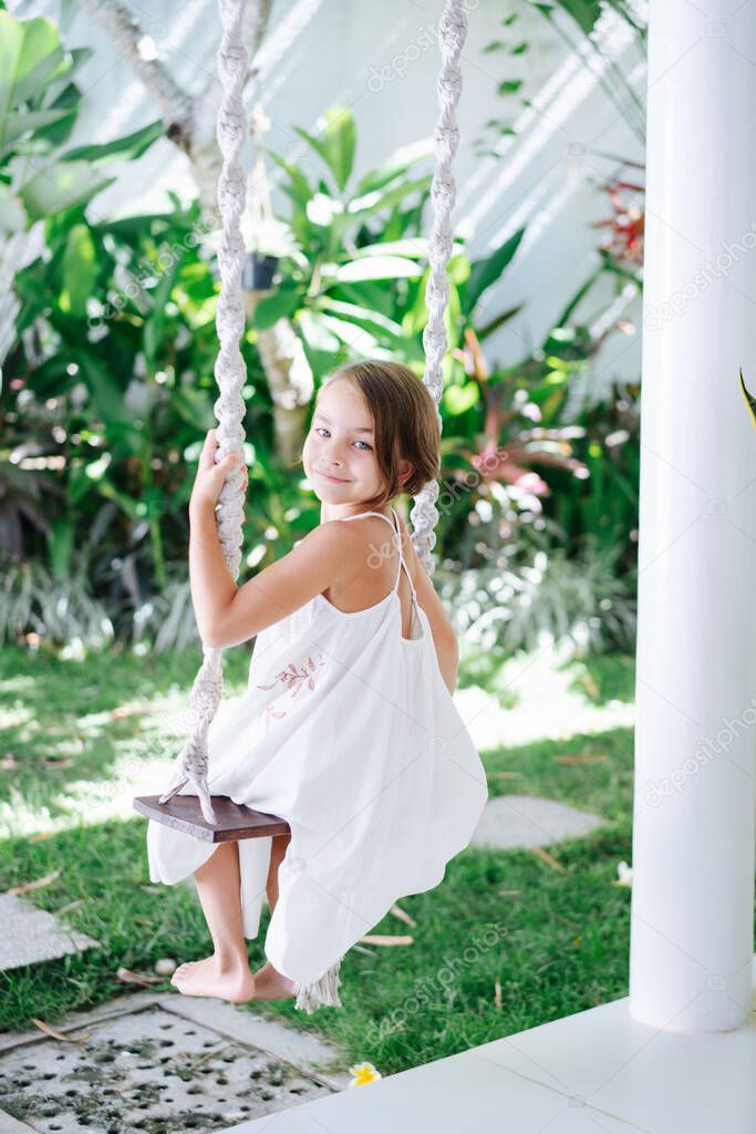 Little brunette girl in a white summer dress sitting on rope swings in a courtyard. From behind. Plants all around. She's looking behind at camera.