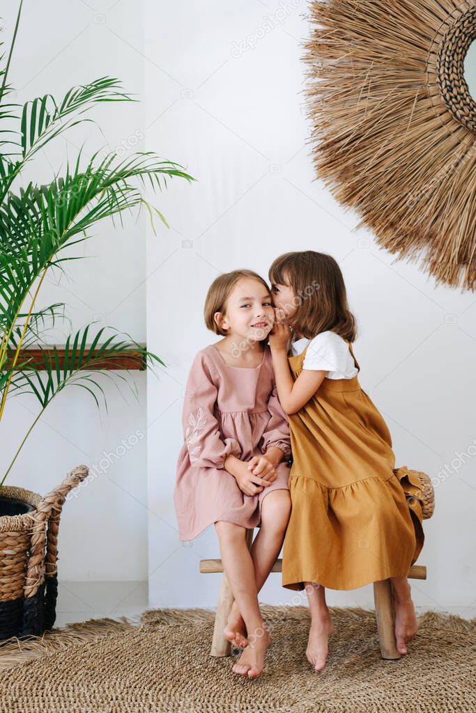 Two little barefoot girls of the same age sitting on a wicker bench in a tropical sttyle room, gossiping. One whispering in the ear, second one looking at camera. Both wearing dresses.