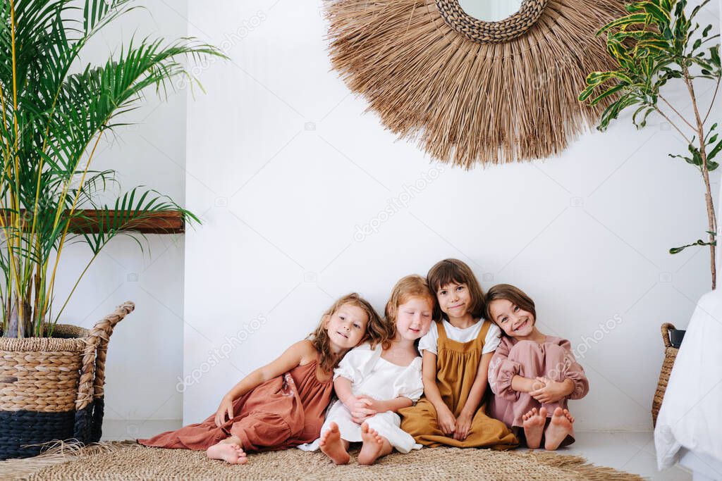 Four happy little girls about the same age are sitting on the floor, leaning on each other, in tropical style room. All wearing dresses and smiling.