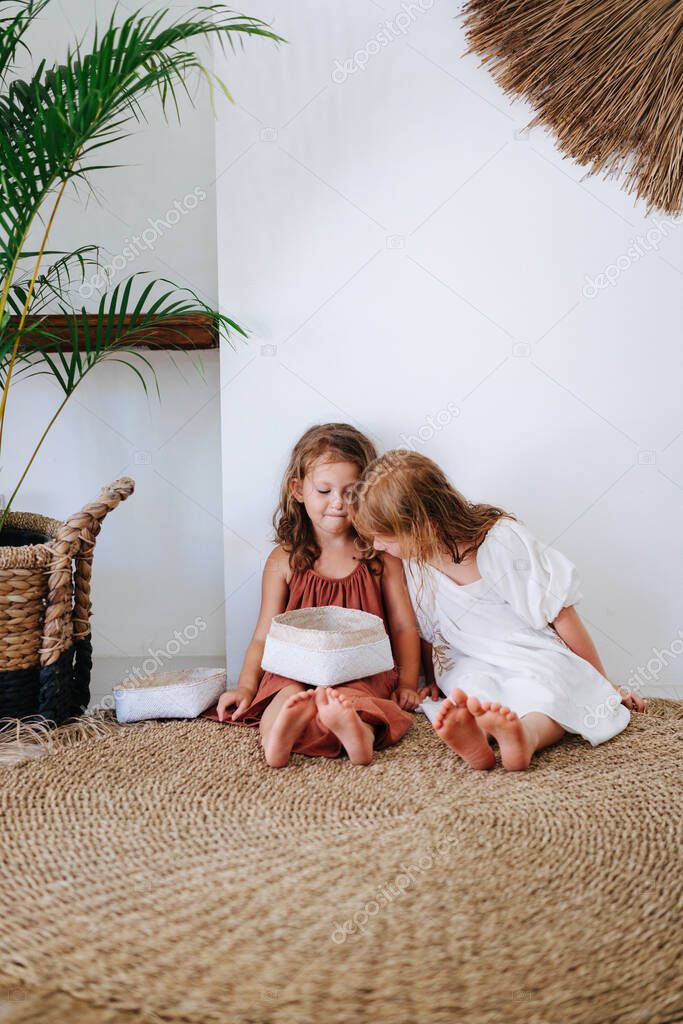 Two curious little girls about the same age are sitting on the floor, in tropical style room, looking inside a box. Both wearing dresses.