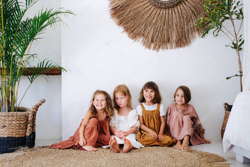 Four happy little girls about the same age are sitting on the floor, shoulder to shoulder, in tropical style room. All wearing dresses and smiling.