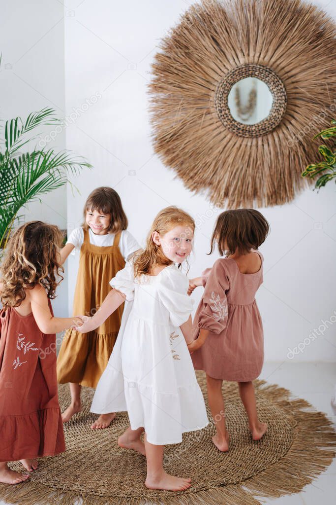 Four happy little girls about the same age walking in a circle with joined hands in tropical style room. Having fun. All barefoot and wearing dresses.