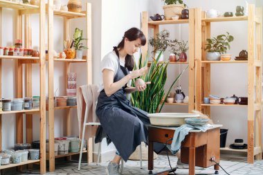 Evdeki atölyede önlüklü esmer kadın çömlekçi çarkına kilden bir top yapıyor.