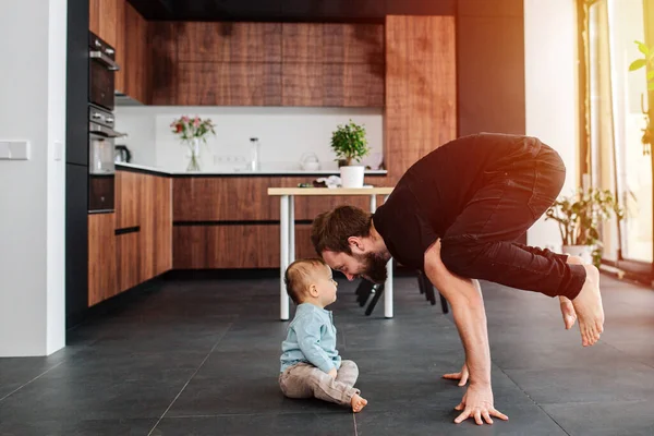 Morning family yoga exercises at home. Father standing on his fingers next to his infant baby. Family quarantine, domestic life in self-isolation. Sunset light from the windows