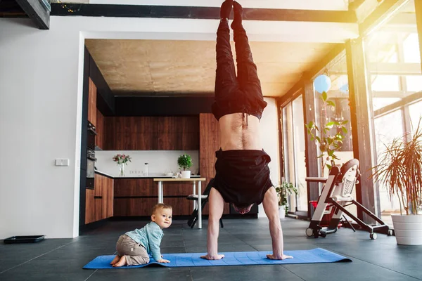 Sports exercises at home, the father performs a handstand, and while the baby crawls on the floor Family quarantine, domestic life in self-isolation.