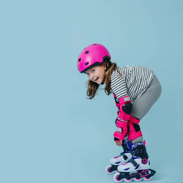 快乐的小女孩 穿着条纹衣服 滑冰鞋 蓝色背景上戴着粉色防护装备 她在卖弄一张照片 保持低矮的安全姿势 — 图库照片