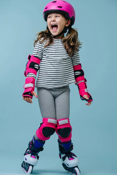 Thrilled happy little girl in striped clothes and roller skates with pink protective gear over blue background. Mouth wide open, knees together, feet apart.