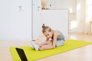 Küçük çocuk evde dizüstü bilgisayarın önünde yoga yapıyor, paspasın üzerinde geriniyor. Evde otururken kendine bakıyor. Sabırlı, ekrana yakından bakıyor..