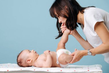 Kundaklama masasında bebek bezi içinde güzel bir bebek yatıyor, genç esmer bir anne onunla oynuyor ve bacağını öpüyor. Mavi bir arkaplanda izole edilmiş