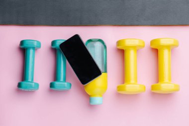 Dört renkli dambıl ve tekrar kullanılabilir plastik su şişesi bir sırada bir paspasın üzerinde duruyor. Üstünde bir ekran olan bir cep telefonu. Üst görünüm.